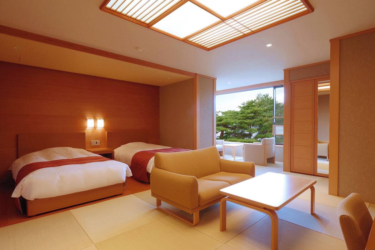 A hotel room with two beds, a sofa, a table, and a view of a tree through large windows.