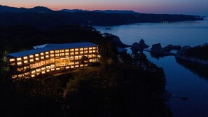 Un edificio de varios pisos con ventanas iluminadas, ubicado cerca de una masa de agua con afloramientos rocosos.