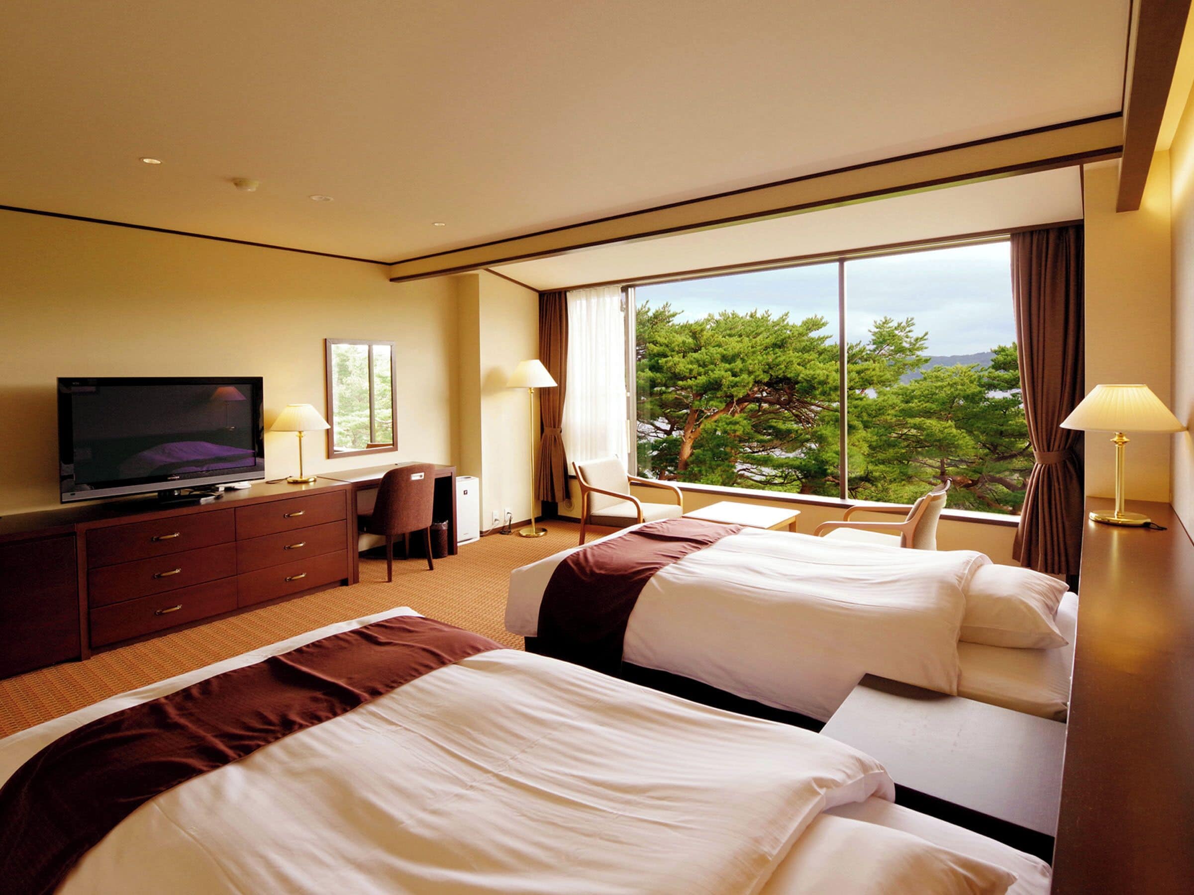 A hotel room with two beds, a desk, a television, and a large window offering a view of trees.