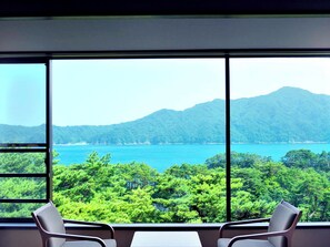 A room with a large window offering a view of a lake and forested hills. There are two chairs and a small table in the room.