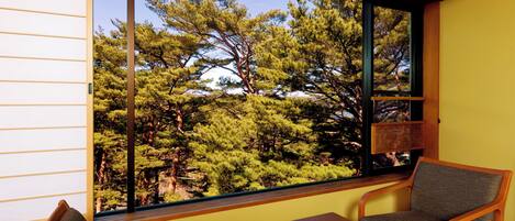 A room with a large window offering a view of pine trees. There are two wooden chairs and a small table.
