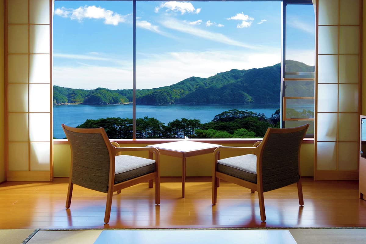 A room with a large window offering a view of a lake and mountains. There are two chairs and a small table in the room.