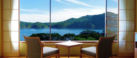 A room with a large window offering a view of a lake and mountains. There are two chairs and a small table in the room.