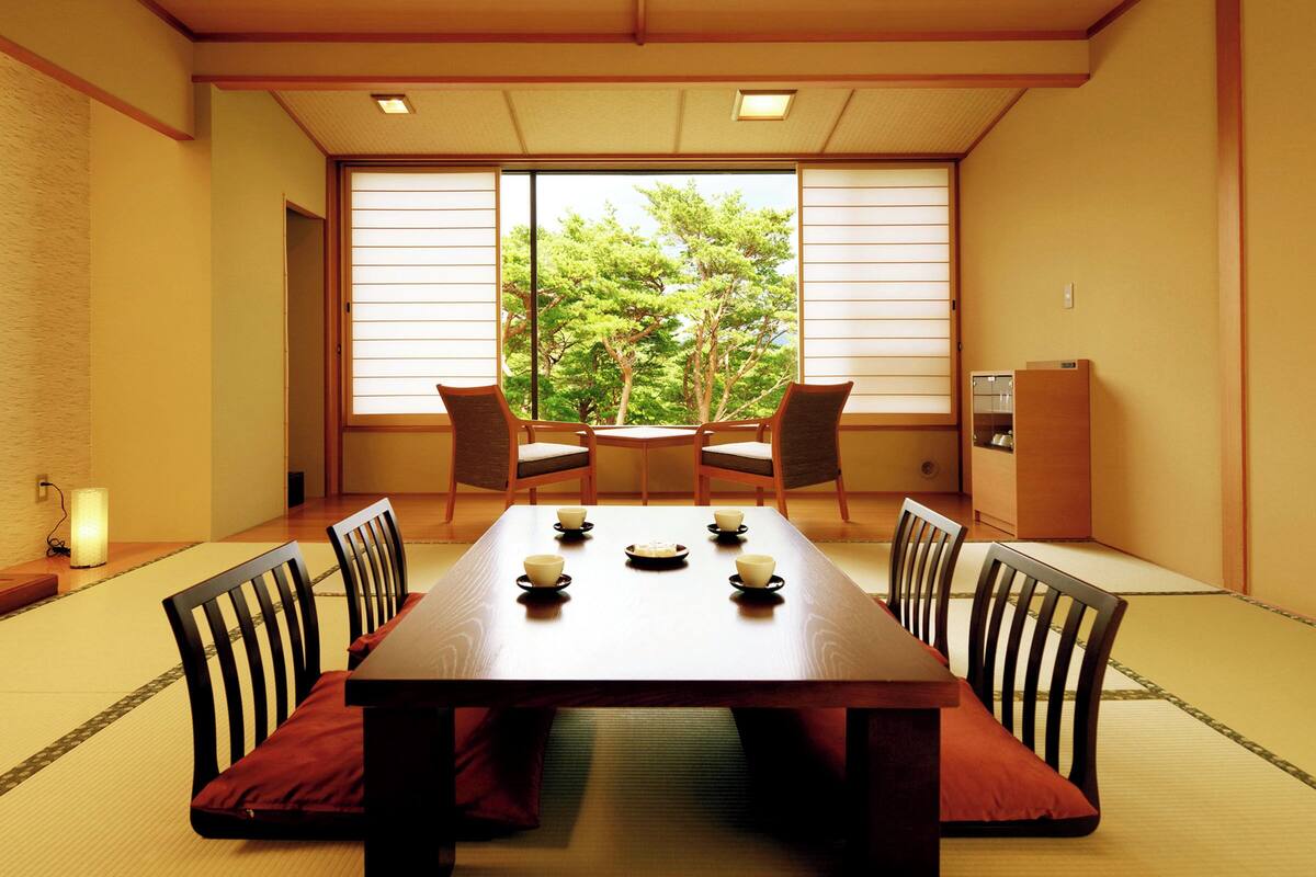 A traditional Japanese room with tatami flooring, a wooden table with cushions, and sliding doors with a view of trees.