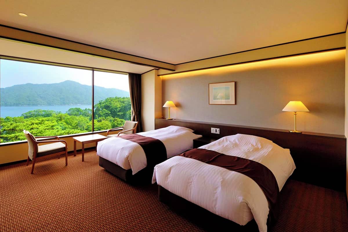 A hotel room with two beds, a desk, and a chair. There is a large window with a view of mountains and greenery.