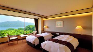 A hotel room with two beds, a desk, and a chair. There is a large window with a view of mountains and greenery.