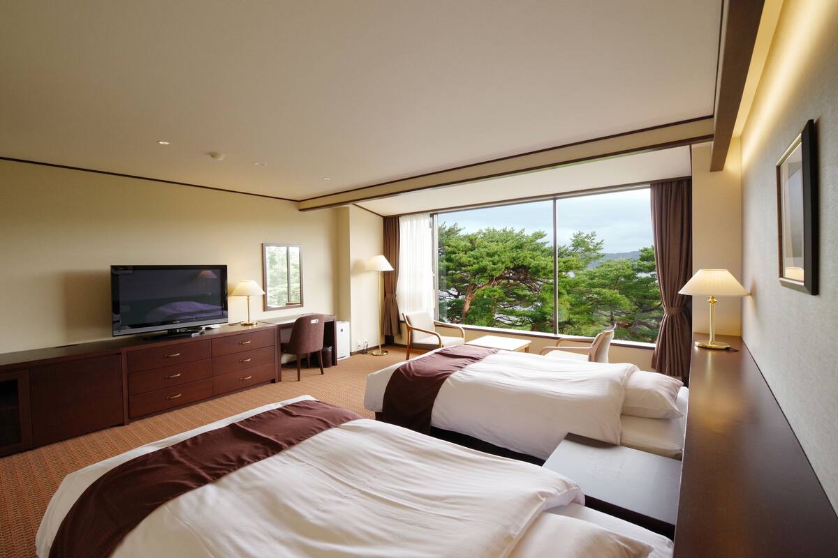 A hotel room with two beds, a desk, a television, and a large window with a view of trees.