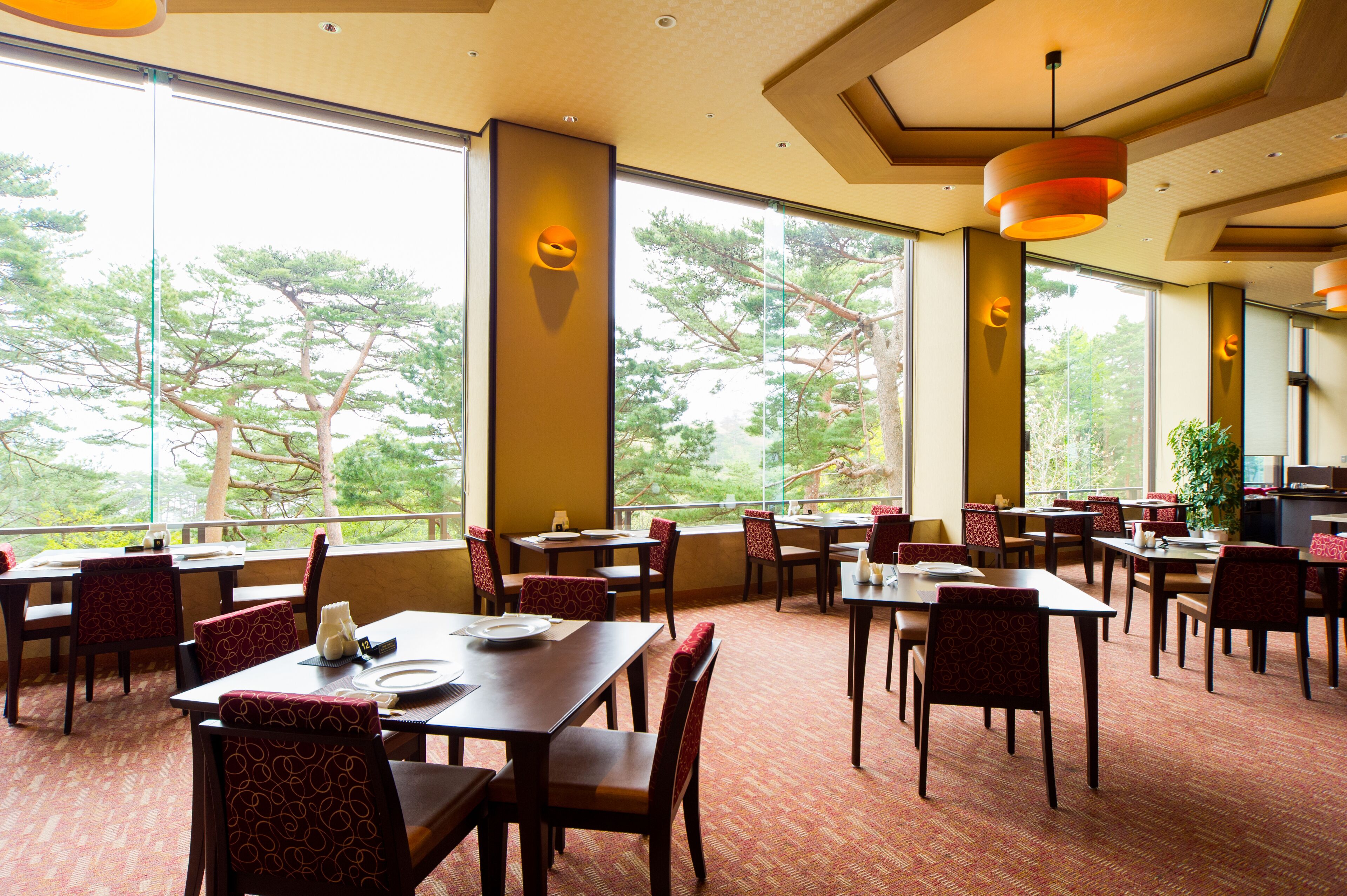 A spacious dining area with multiple tables and chairs, large windows offering a view of trees, and modern pendant lighting.