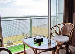 A balcony with wicker chairs and a glass table, overlooking a body of water and a distant shoreline.