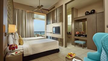 A hotel room with a bed, a desk with a lamp, a TV, a dining table with fruit, a blue armchair, and a view of the sea.