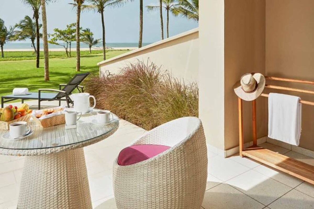 A balcony with a glass table, wicker chairs, a hat rack with a towel, and a view of the beach and palm trees.