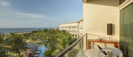 A balcony with a glass railing overlooking a swimming pool, palm trees, and a view of the ocean.