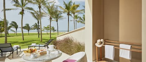 A balcony with wicker furniture, a table with breakfast items, and a view of the ocean and palm trees.