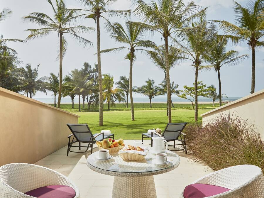 Ein Balkon mit Korbstühlen, einem Glastisch mit Frühstücksgebäck und Blick auf Palmen und den Strand.