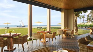 An outdoor dining area with tables and chairs, overlooking a beach with umbrellas and a sailboat. - Sentido Heritance Negombo (Negombo)