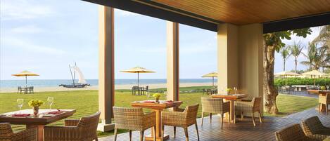 An outdoor dining area with tables and chairs, overlooking a beach with umbrellas and a sailboat.