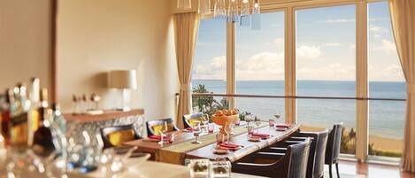 A dining area with a table set for six, large windows offering a view of the ocean, and hanging pendant lights.