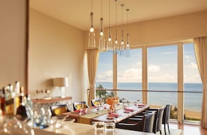 A dining area with a table set for six, large windows offering a view of the ocean, and hanging pendant lights.