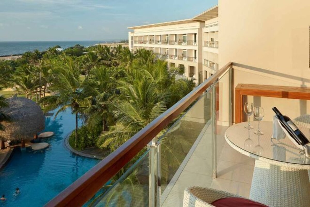 Un balcón con barandilla de vidrio que da a una piscina rodeada de palmeras, una estructura con techo de paja y un edificio de hotel.