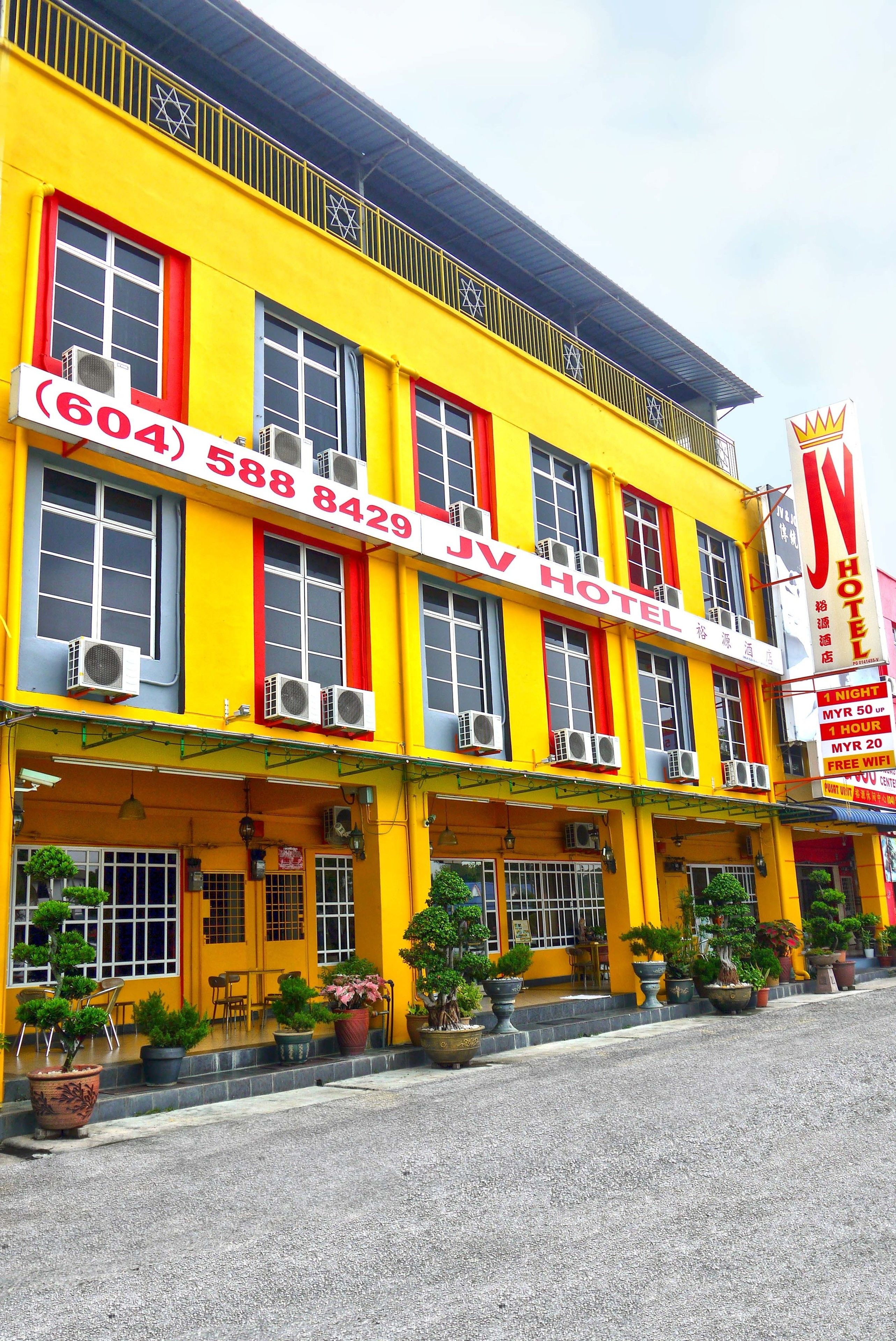 A hotel with a bright yellow facade, multiple air conditioning units, and a signboard with contact numbers.