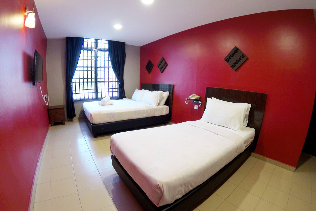 Habitación de hotel con dos camas, una ventana con cortinas, un teléfono sobre un soporte y una pared roja con una placa.