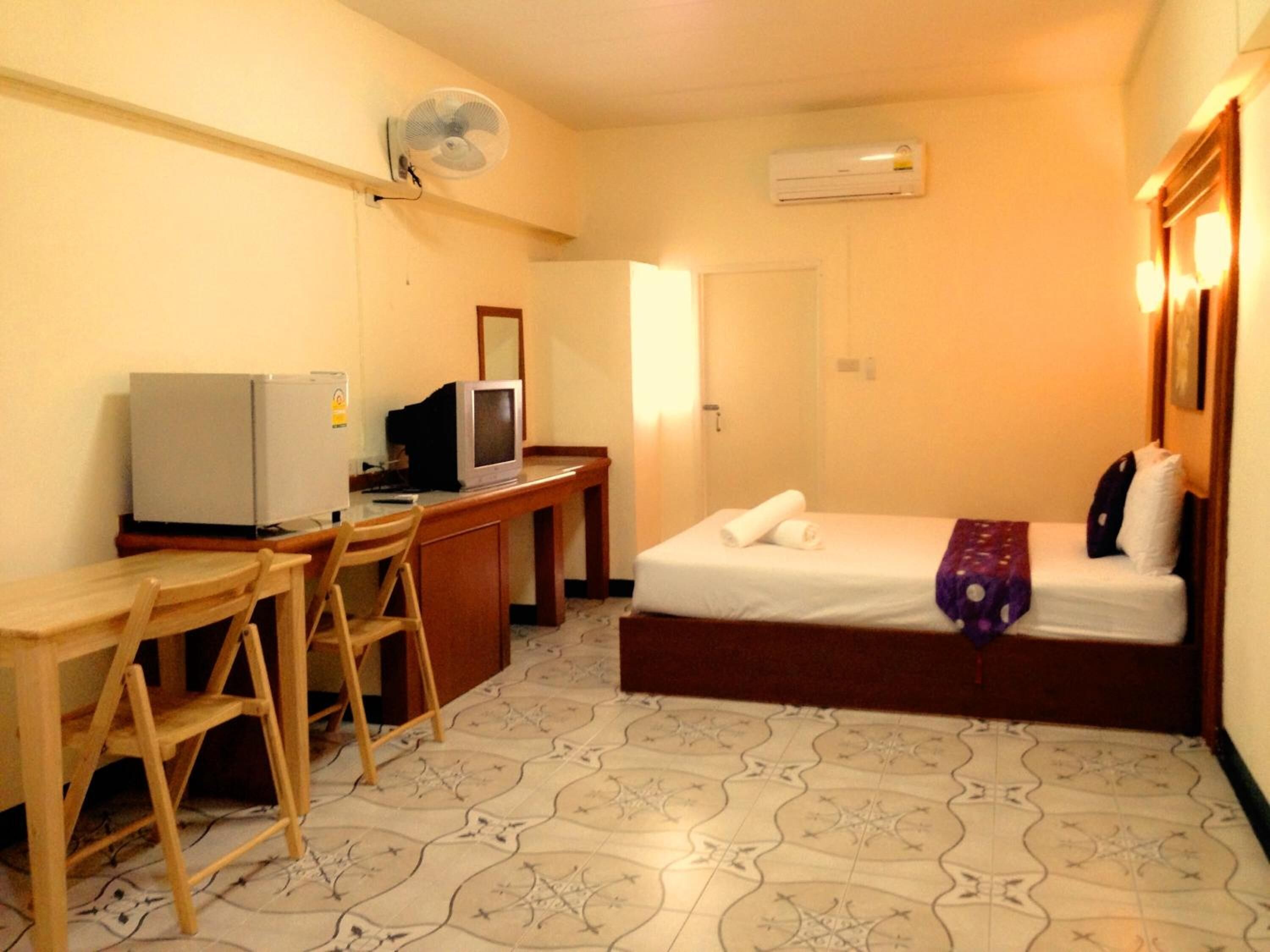Una habitación de hotel pequeña con una cama, un escritorio con computadora y televisor, una silla y un ventilador.