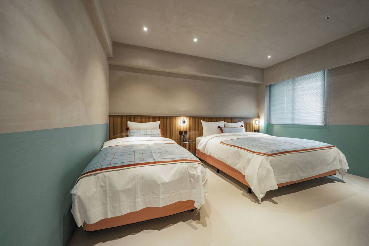 Una habitación de hotel con dos camas, un cabecero de madera, un ventanal con persianas y una lámpara fijada a la pared.