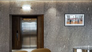 Um elevador com acabamento metálico, uma moldura com um quadro na parede e uma mesa redonda metálica.