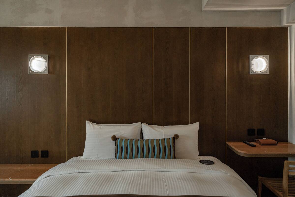 Una habitación de hotel con cabecera de madera, cama con ropa blanca, almohada a rayas, mesita de noche con un control remoto y una silla.