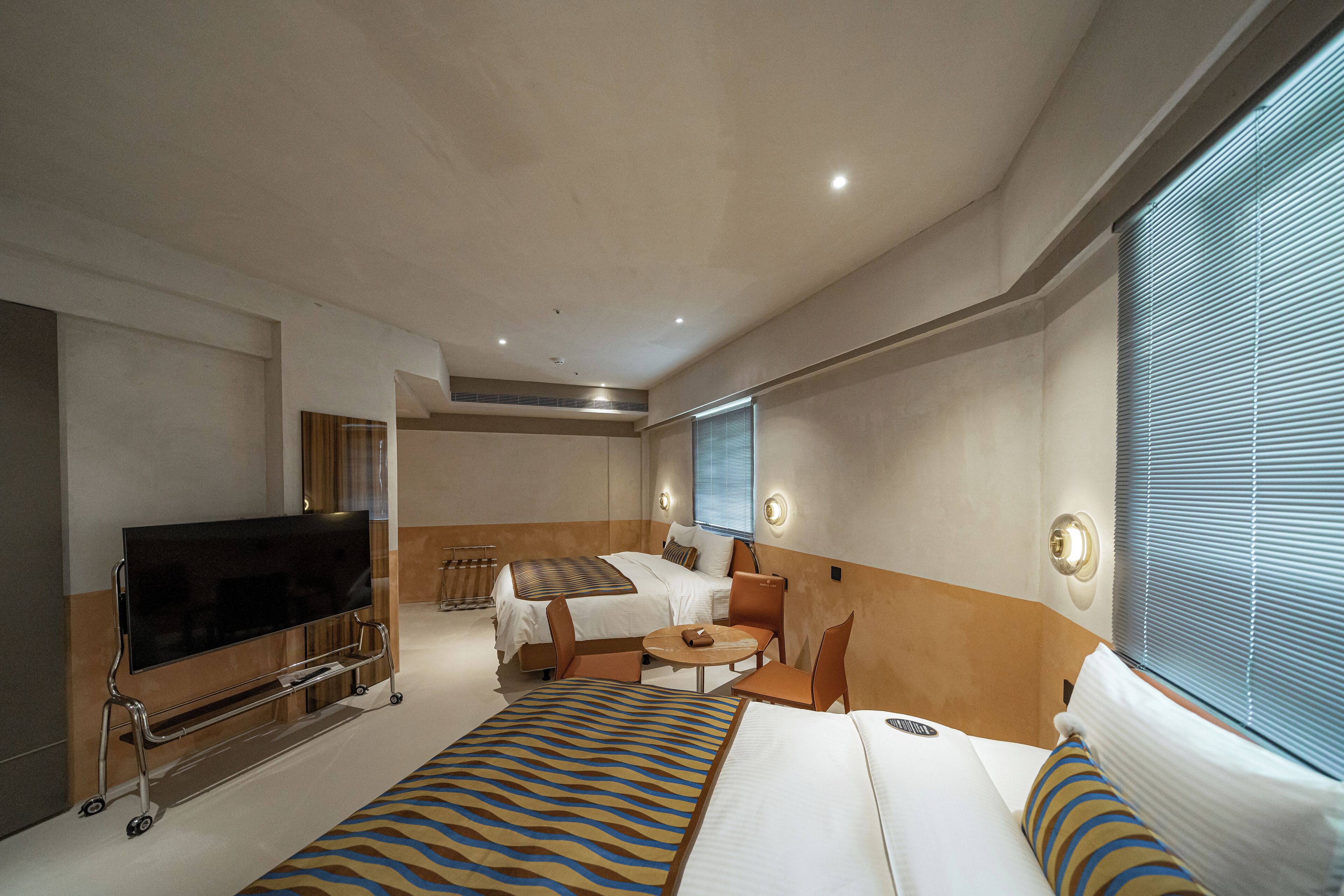 A hotel room with a bed, a TV on a stand, a small table, and a window with blinds.