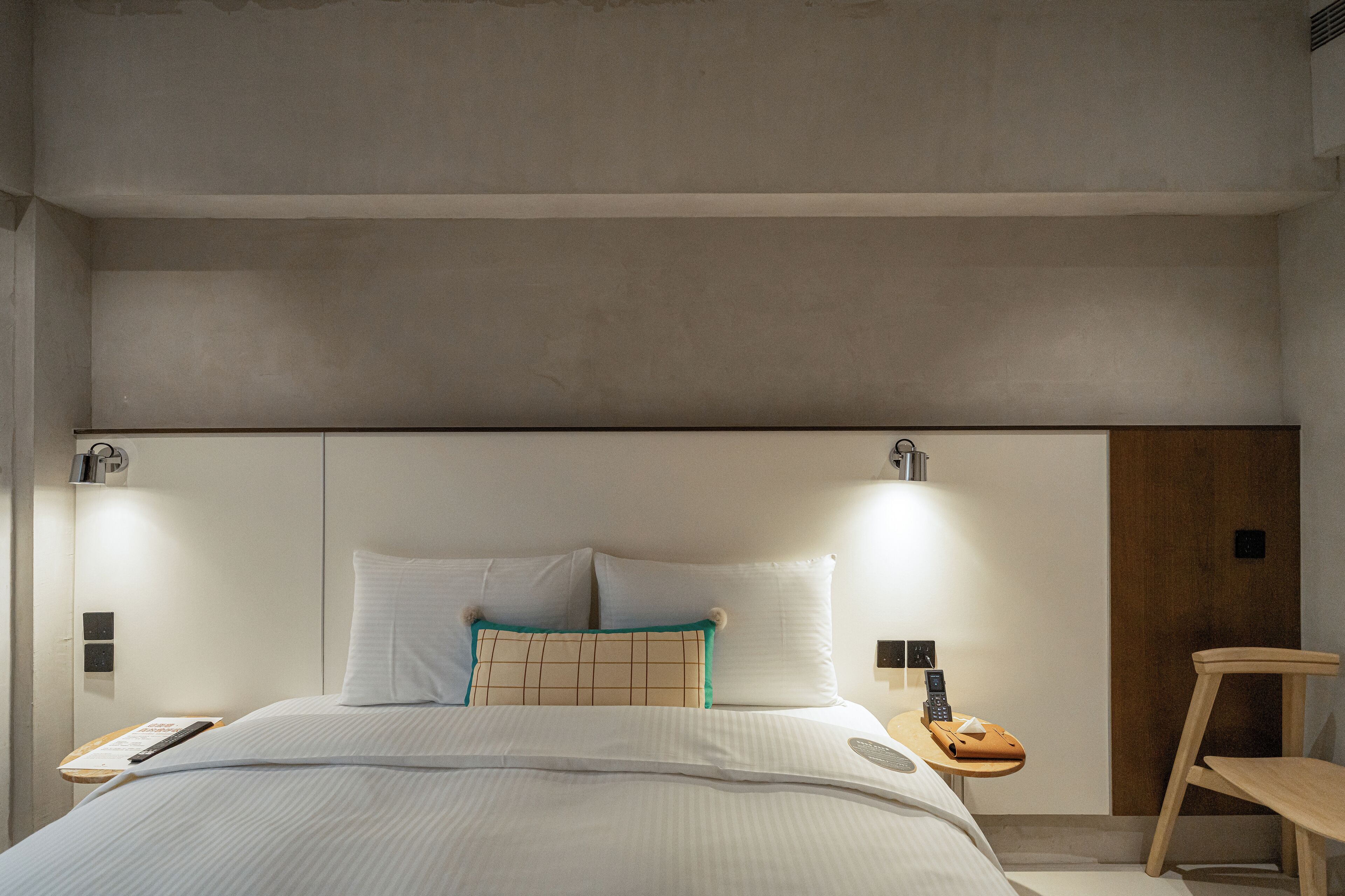A modern hotel room with a large bed, bedside tables, a wooden chair, and a nightstand with a phone and remote control.