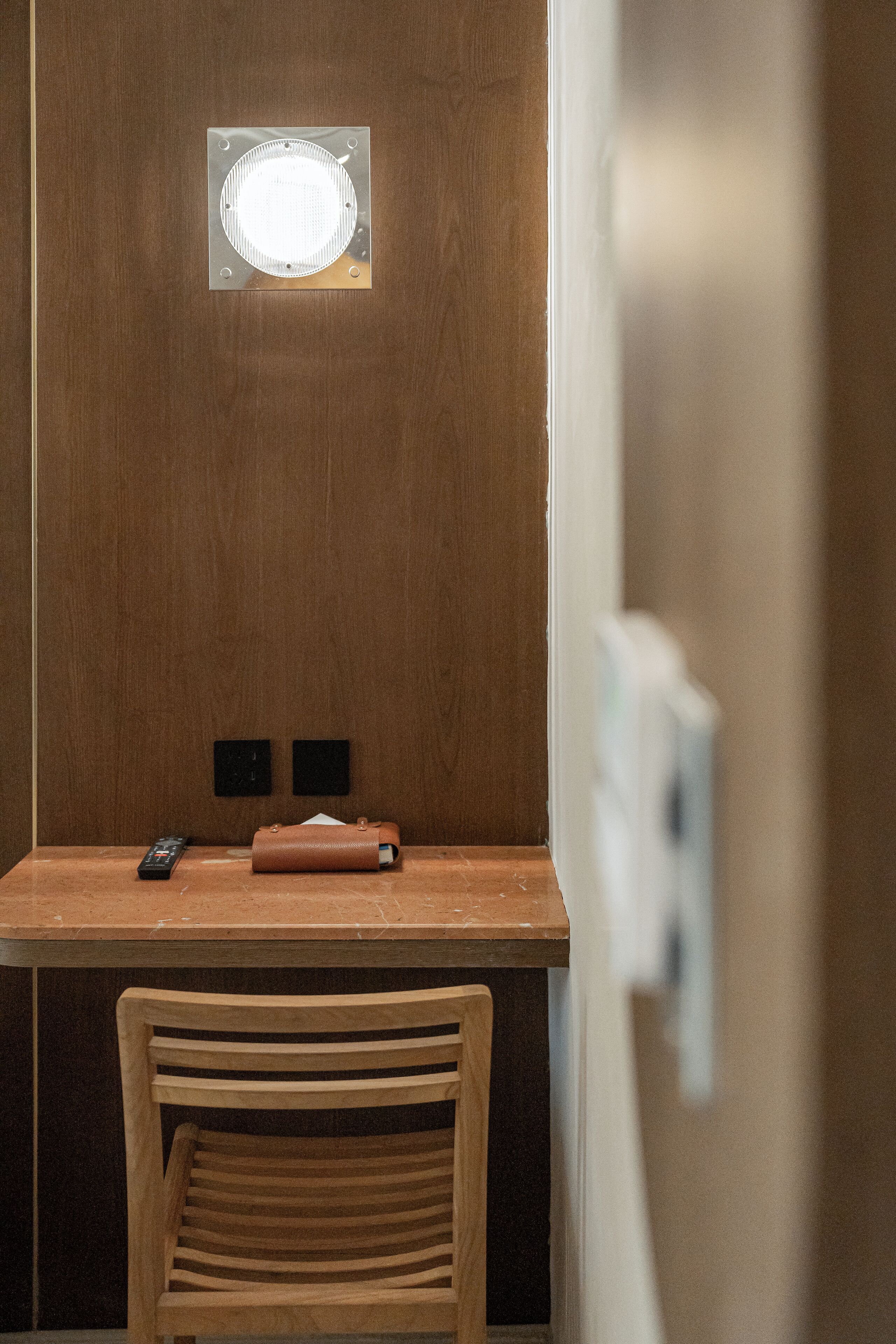 A wooden desk with a remote control and a leather bag, a wooden chair, and a wall-mounted light fixture.