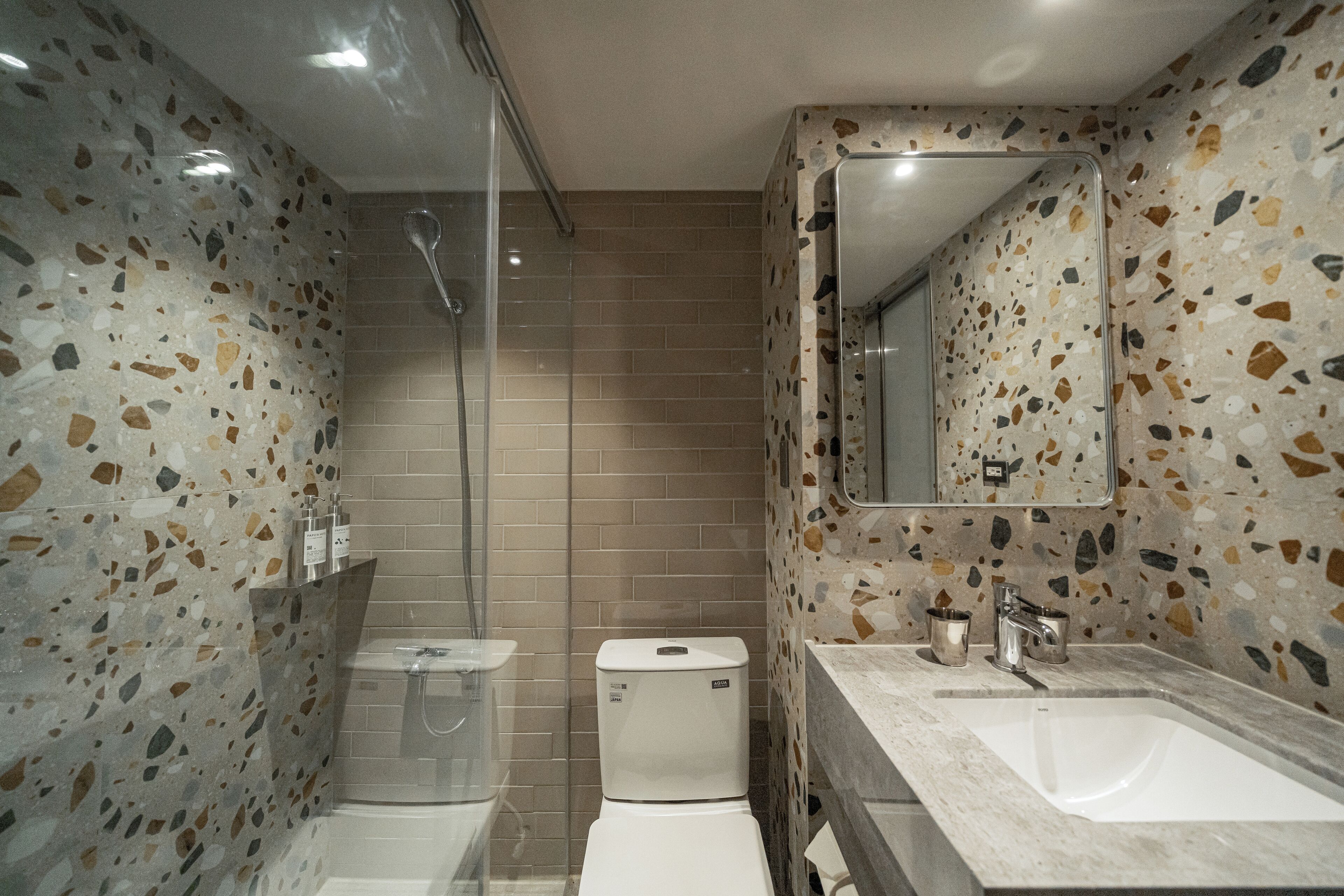 A bathroom with a shower, toilet, and sink, featuring terrazzo flooring and a mirror above the sink.