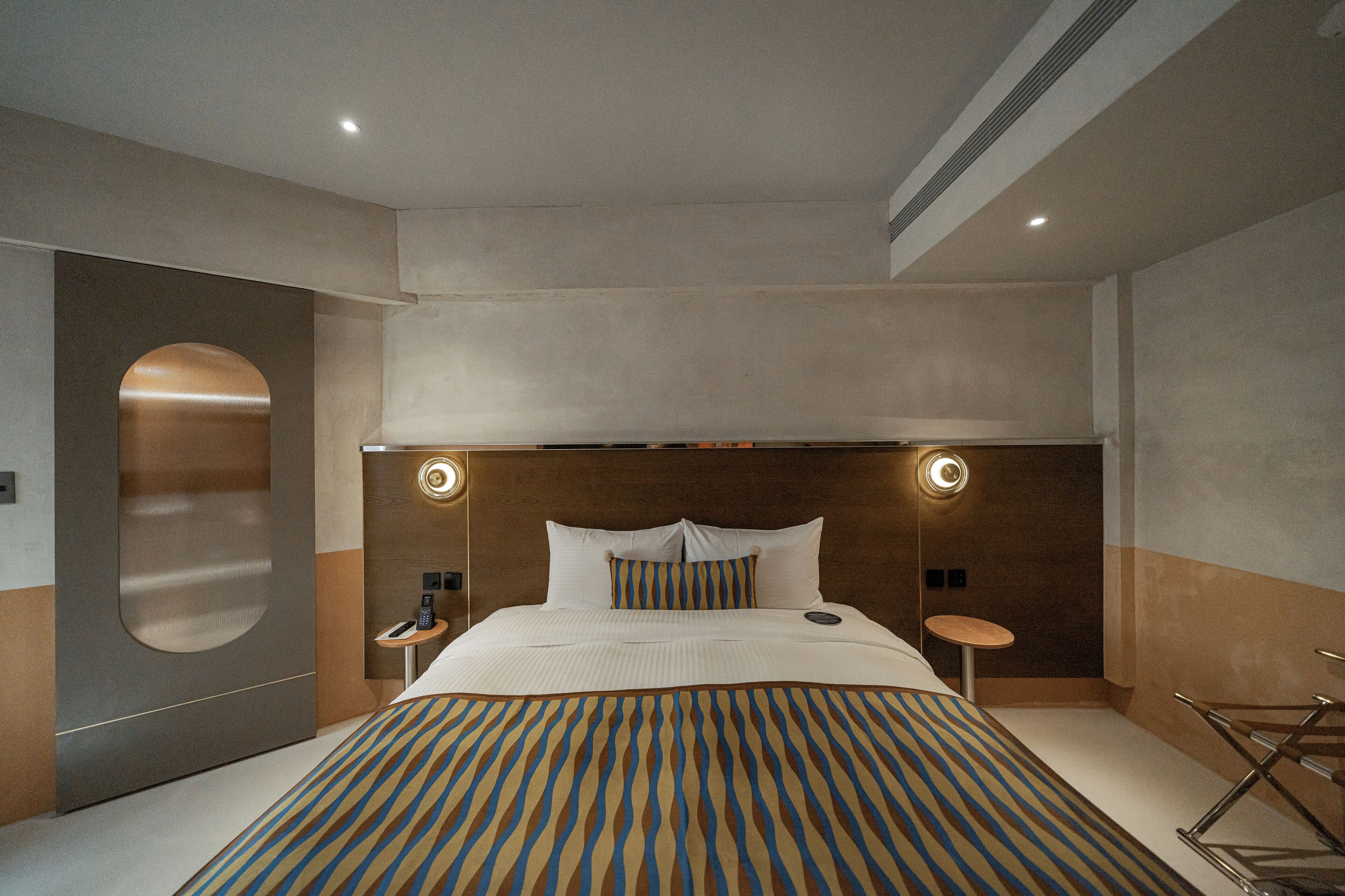 Una habitación de hotel moderna con una cama grande, dos mesitas de noche, un espejo redondo y un armario empotrado.