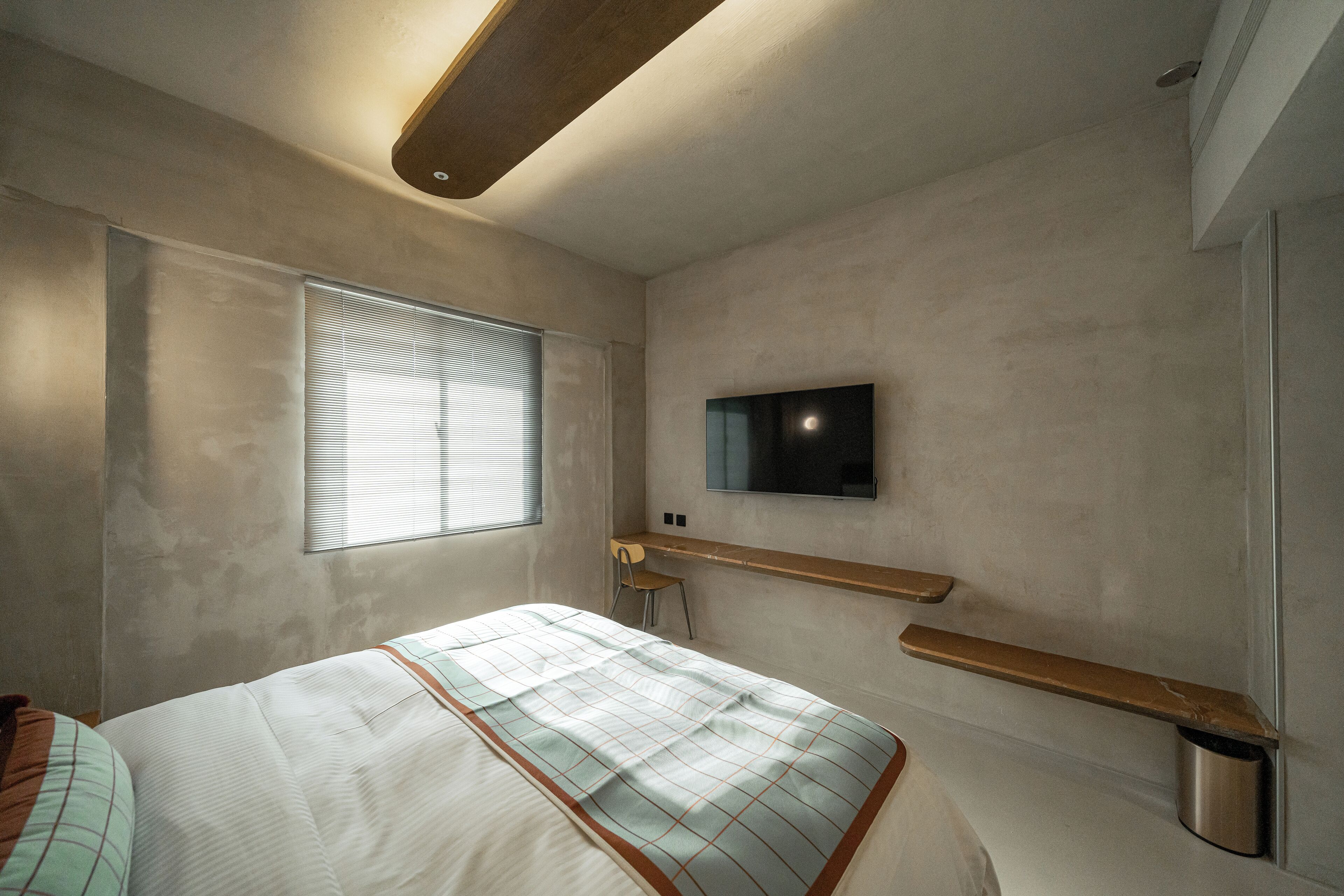 A modern bedroom with a bed, a TV mounted on the wall, a window with blinds, and a wooden shelf.