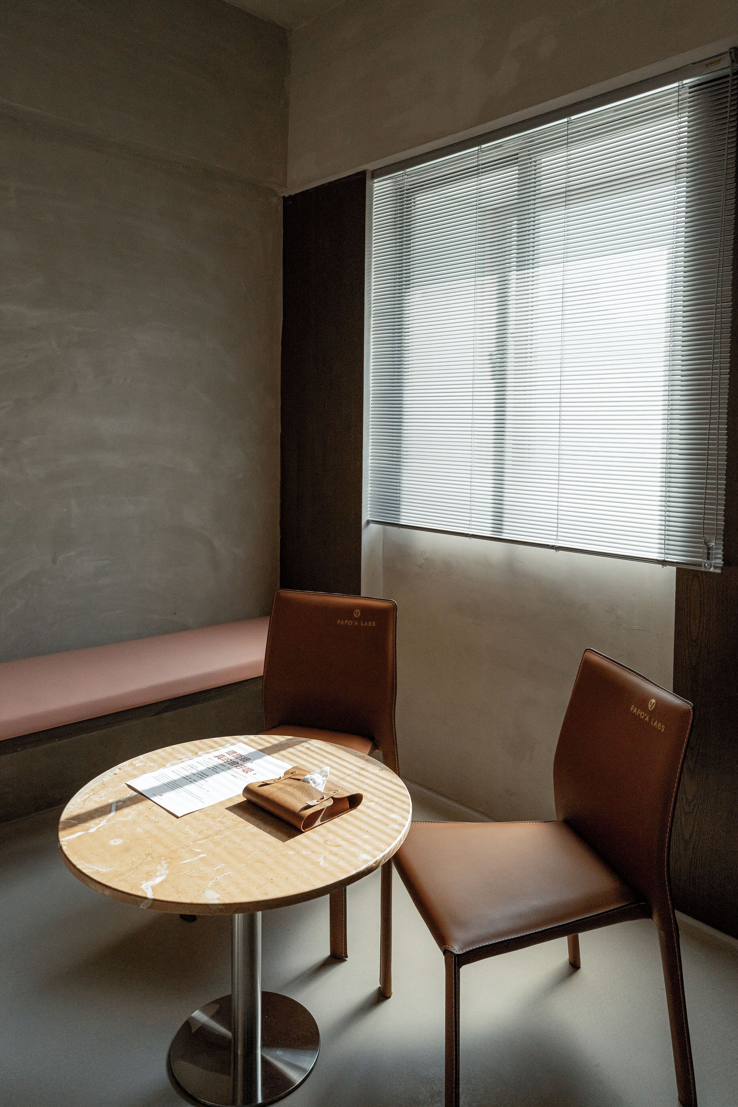 A room with a round table, two chairs, a bench, and a window with blinds.