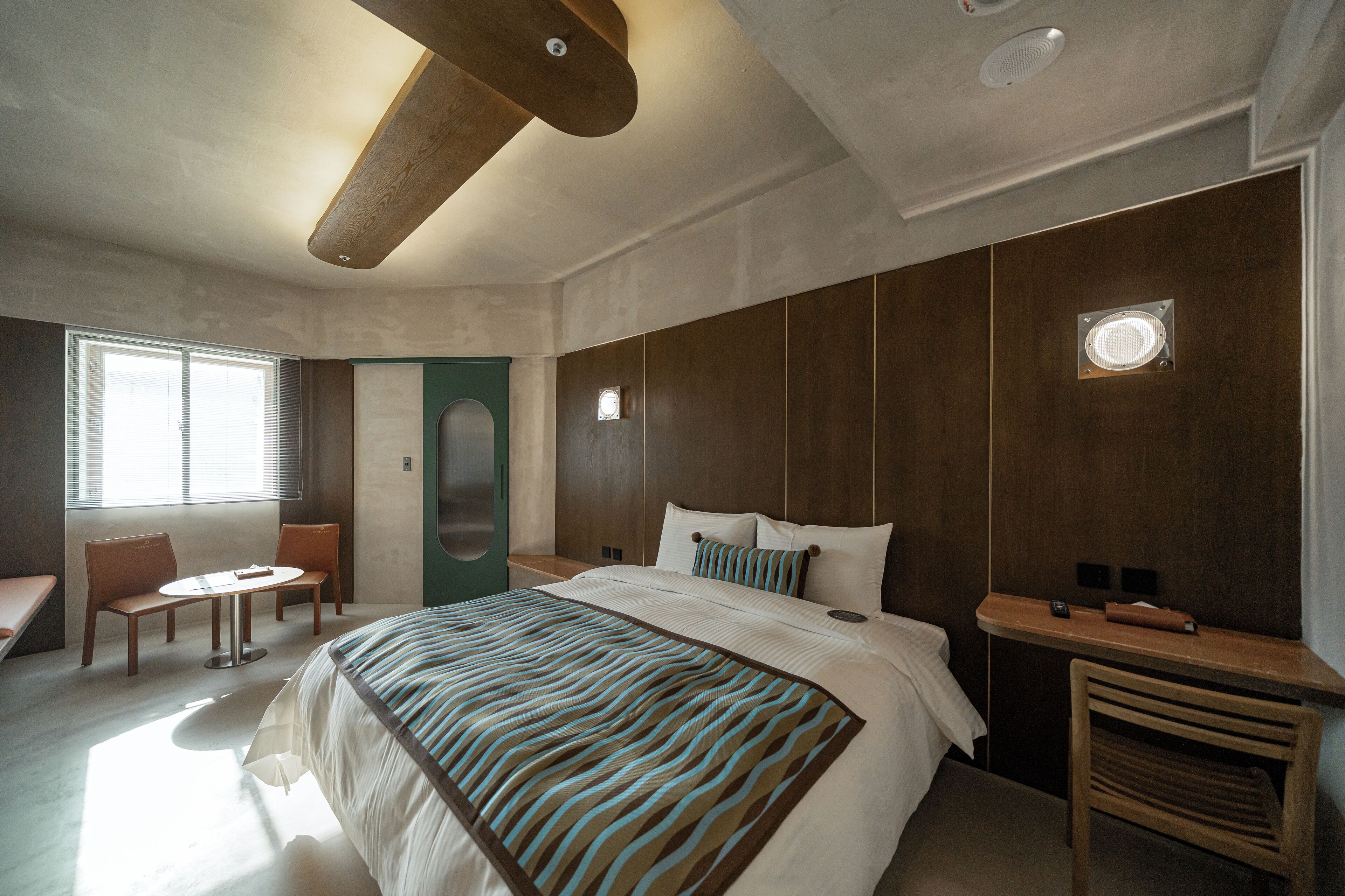 A modern hotel room with a large bed, a small round table, two chairs, a wooden bench, and a built-in desk with a lamp.