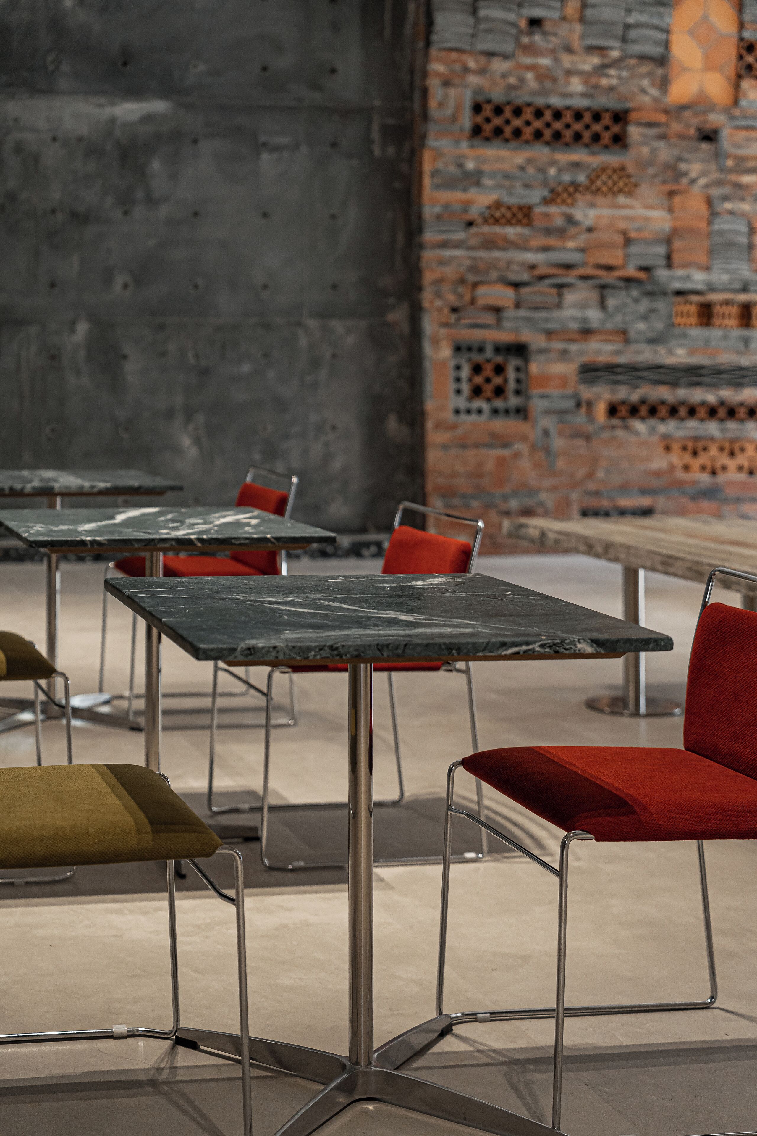 A modern interior with metal-framed tables and red upholstered chairs, set against a textured brick wall.