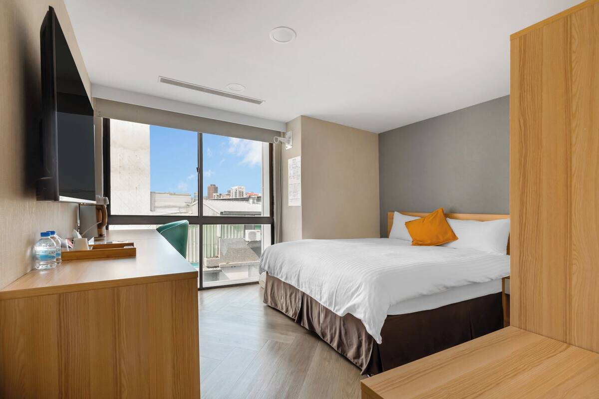A modern hotel room with a bed, a desk, a television, and a large window offering a city view.