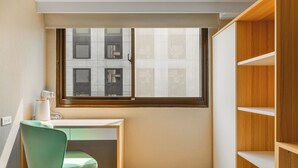Una habitación pequeña y bien iluminada con un escritorio, una silla verde, una ventana con persianas y estanterías de madera empotradas.