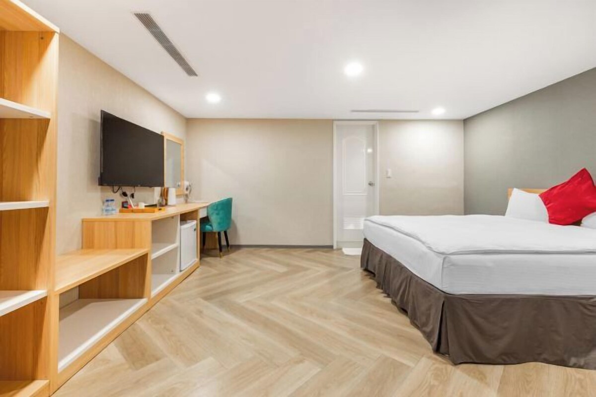 A modern hotel room with a bed, a desk, a chair, a TV, and built-in shelving.