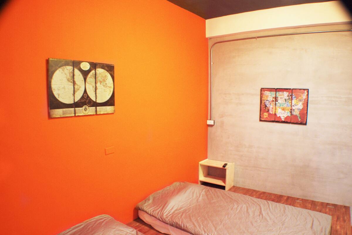 A room with orange walls, a bed with two pillows, a small wooden nightstand, and two wall-mounted artworks.