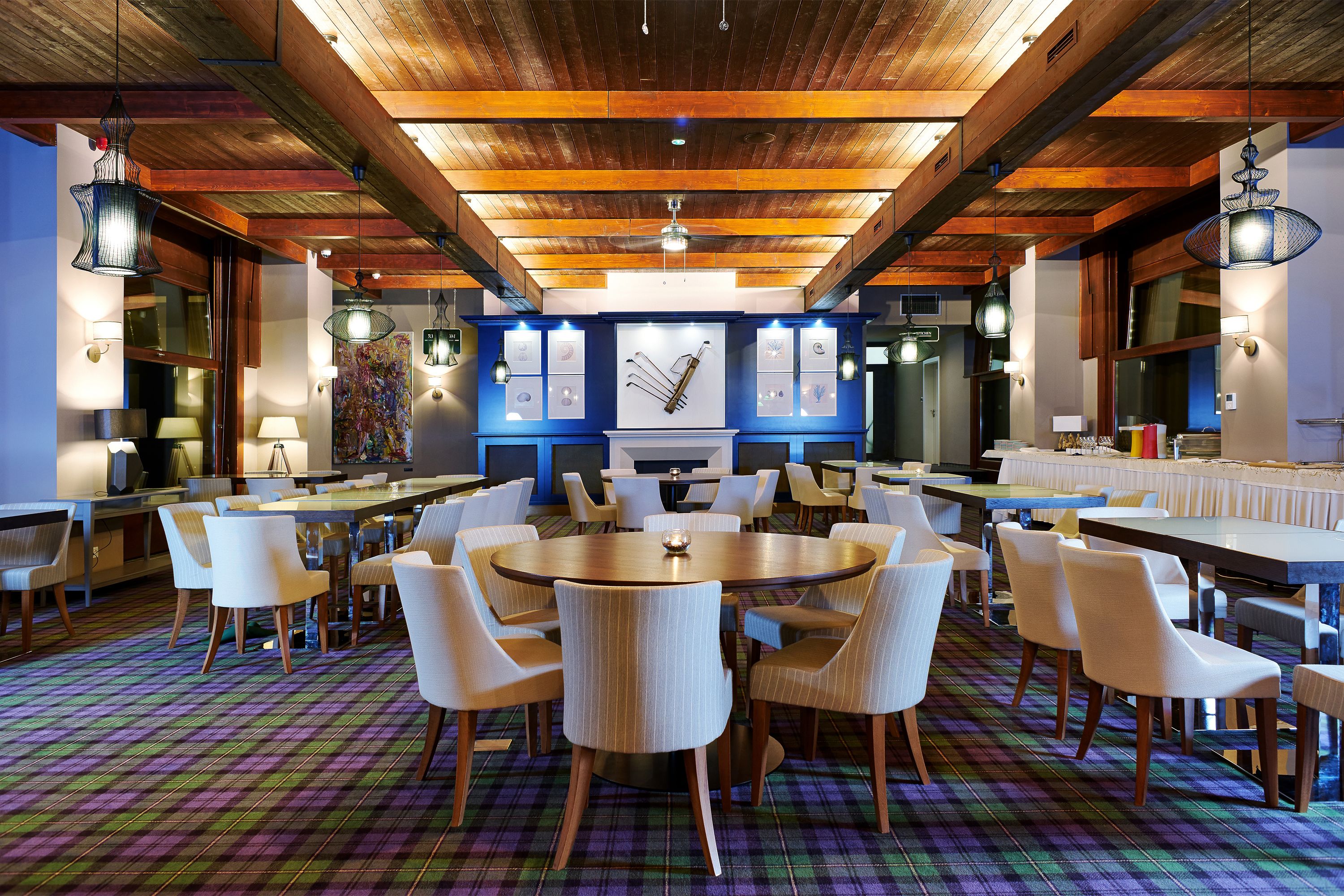 A spacious dining area with round tables and white chairs, wooden beams on the ceiling, and a colorful plaid carpet.