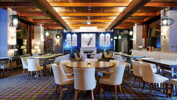 A spacious dining area with round tables and white chairs, wooden beams on the ceiling, and a colorful plaid carpet.