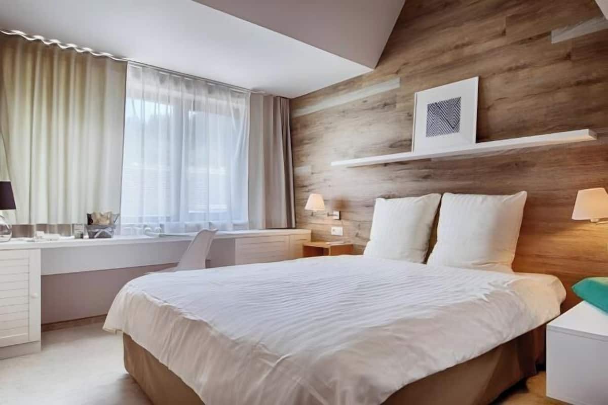 Een slaapkamer met een bed, een bureau, een stoel, een raam met gordijnen, en een wandplank met een foto.