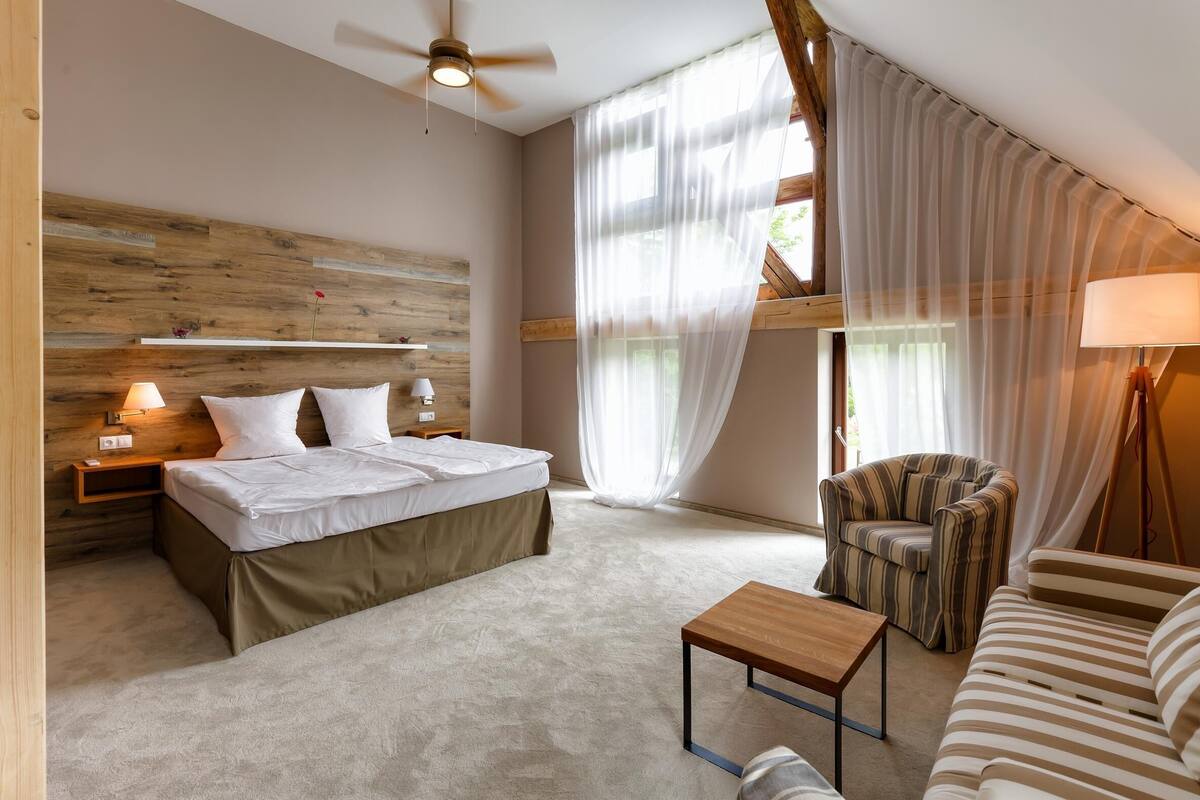 Een slaapkamer met een bed, nachtlampen, een houten hoofdbord, een plafondventilator, een raam met dunne gordijnen, een gestreept fauteuil, een gestreepte bank en een klein houten tafeltje.