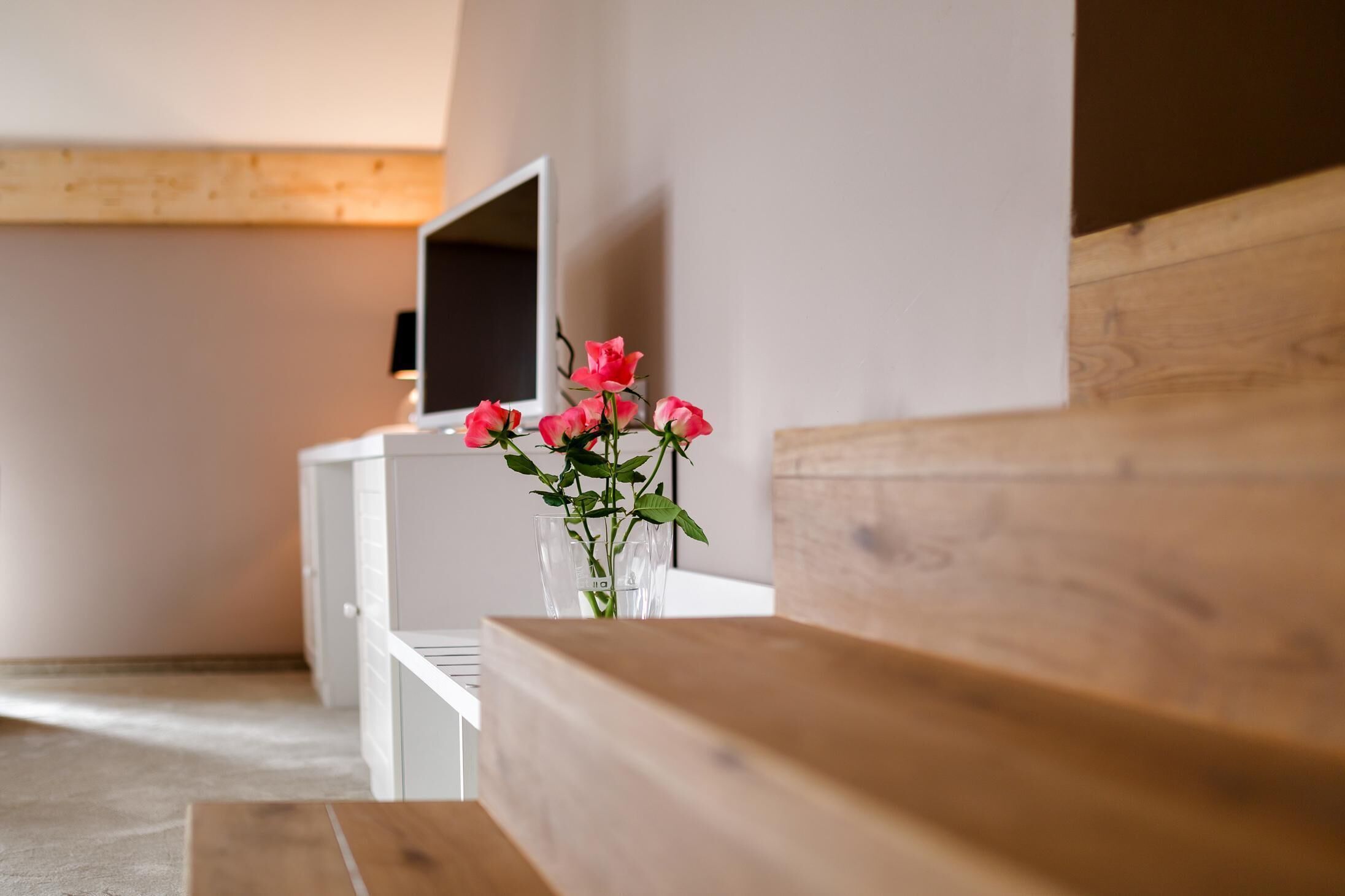 Una stanza con una panca in legno, una scrivania bianca e un vaso con fiori rosa.