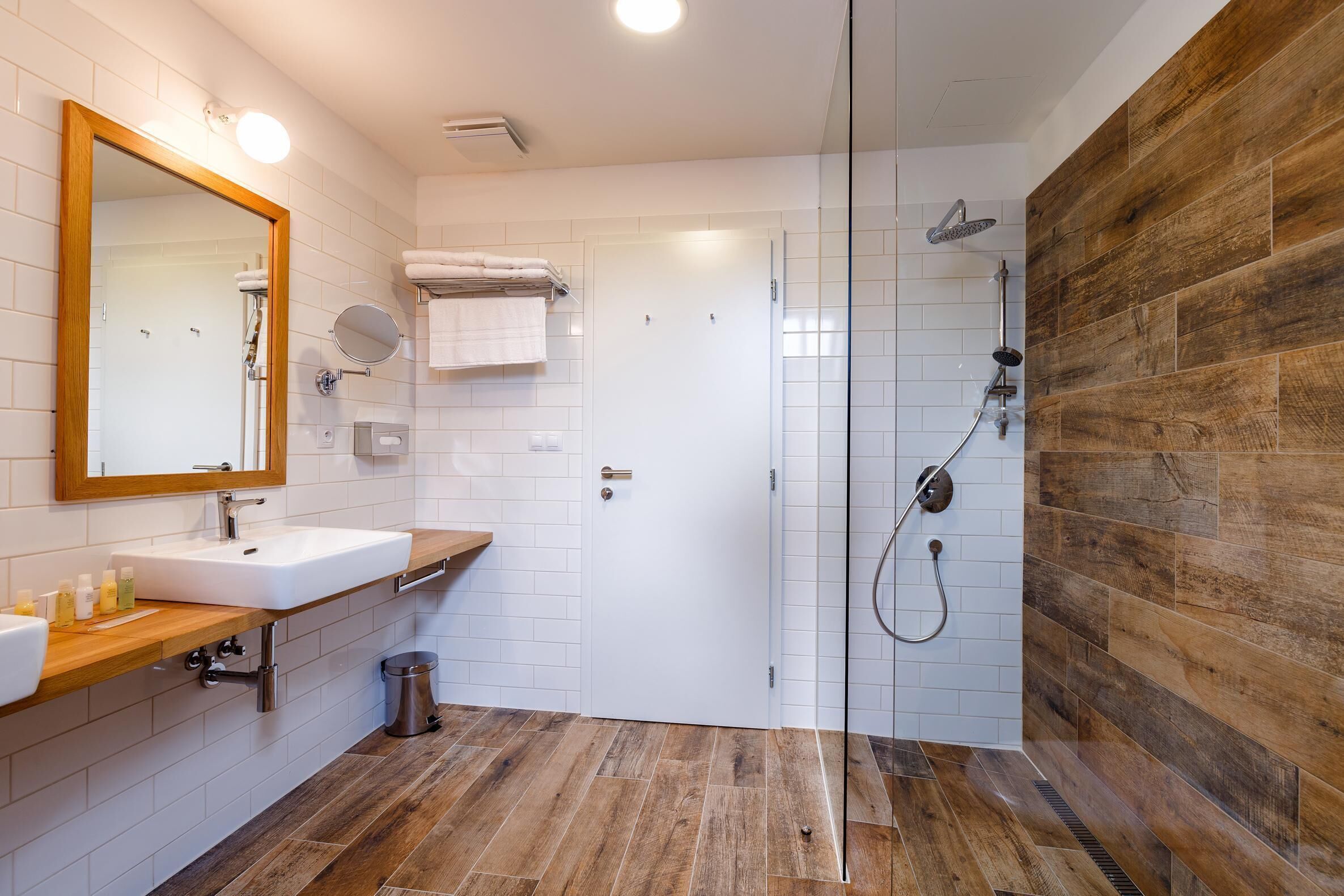 Un baño moderno con piso de madera, azulejos blancos, un espejo grande, un lavamanos y una zona de ducha.