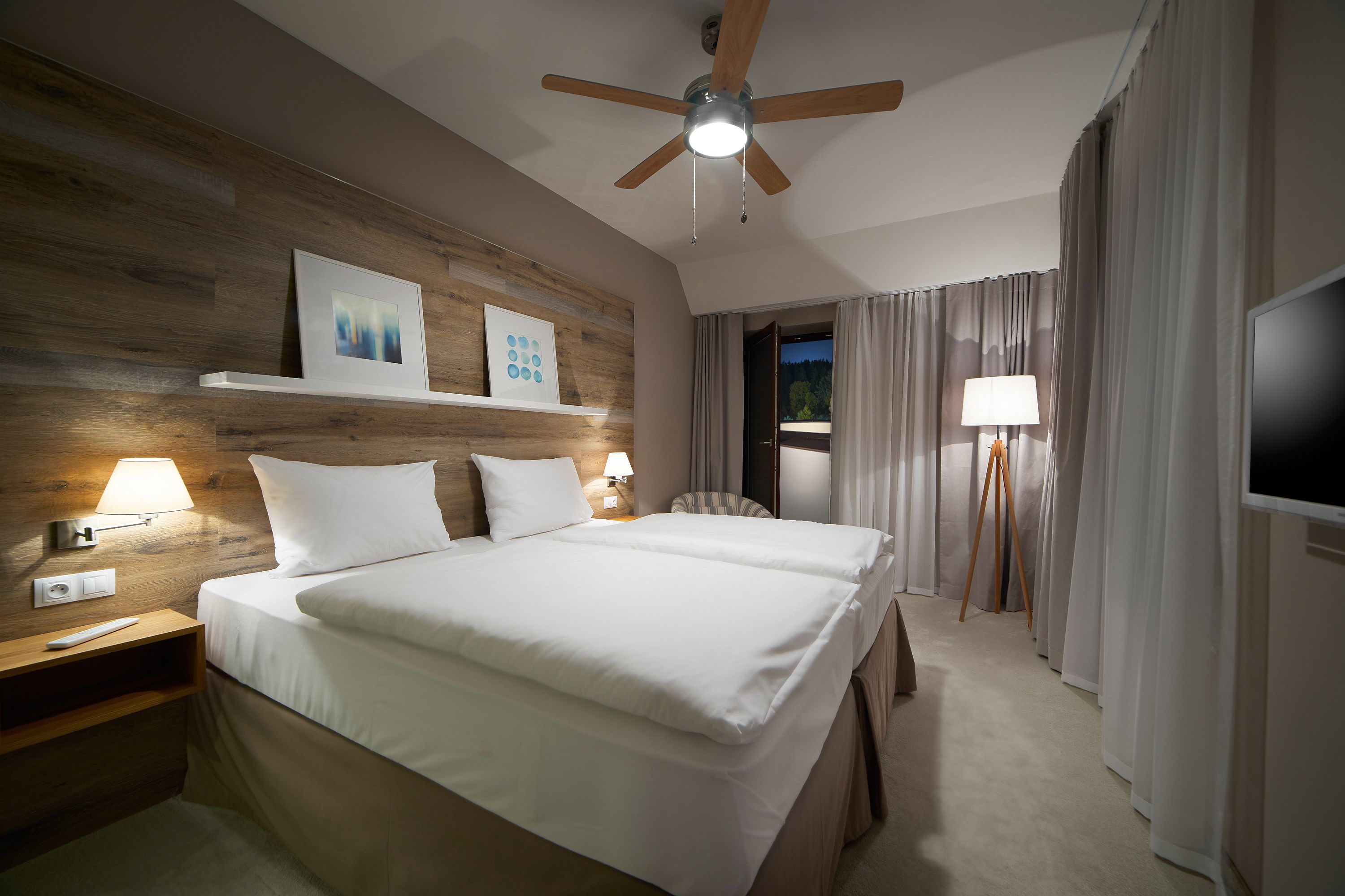 A hotel room with a large bed, a ceiling fan, a TV, and a nightstand with a lamp.