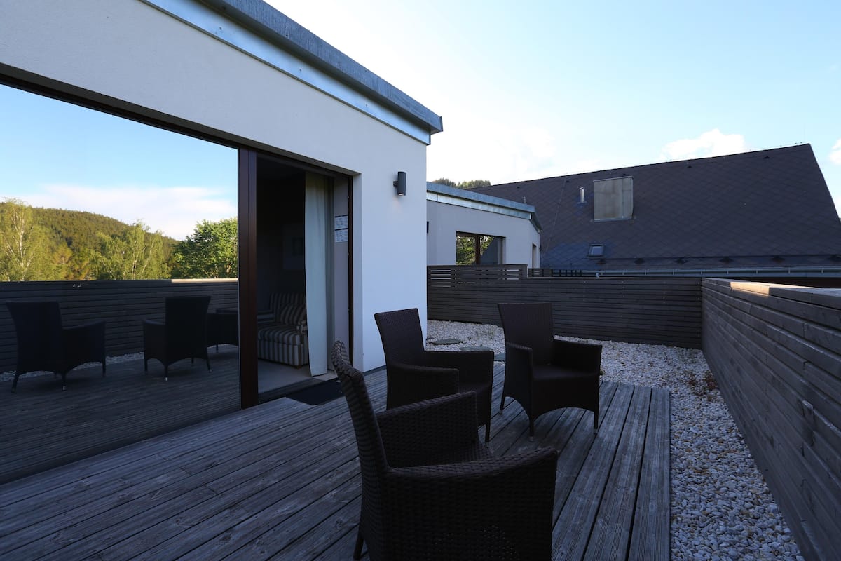Een modern terras buiten met wicker meubilair, een houten vloer en een gebouw met een donker dak.